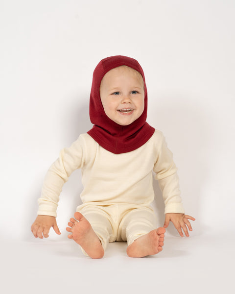 A toddler girl sitting on the ground, wearing the menique kids' merino 160gsm balaclava in royal cherry color, studio background picture.