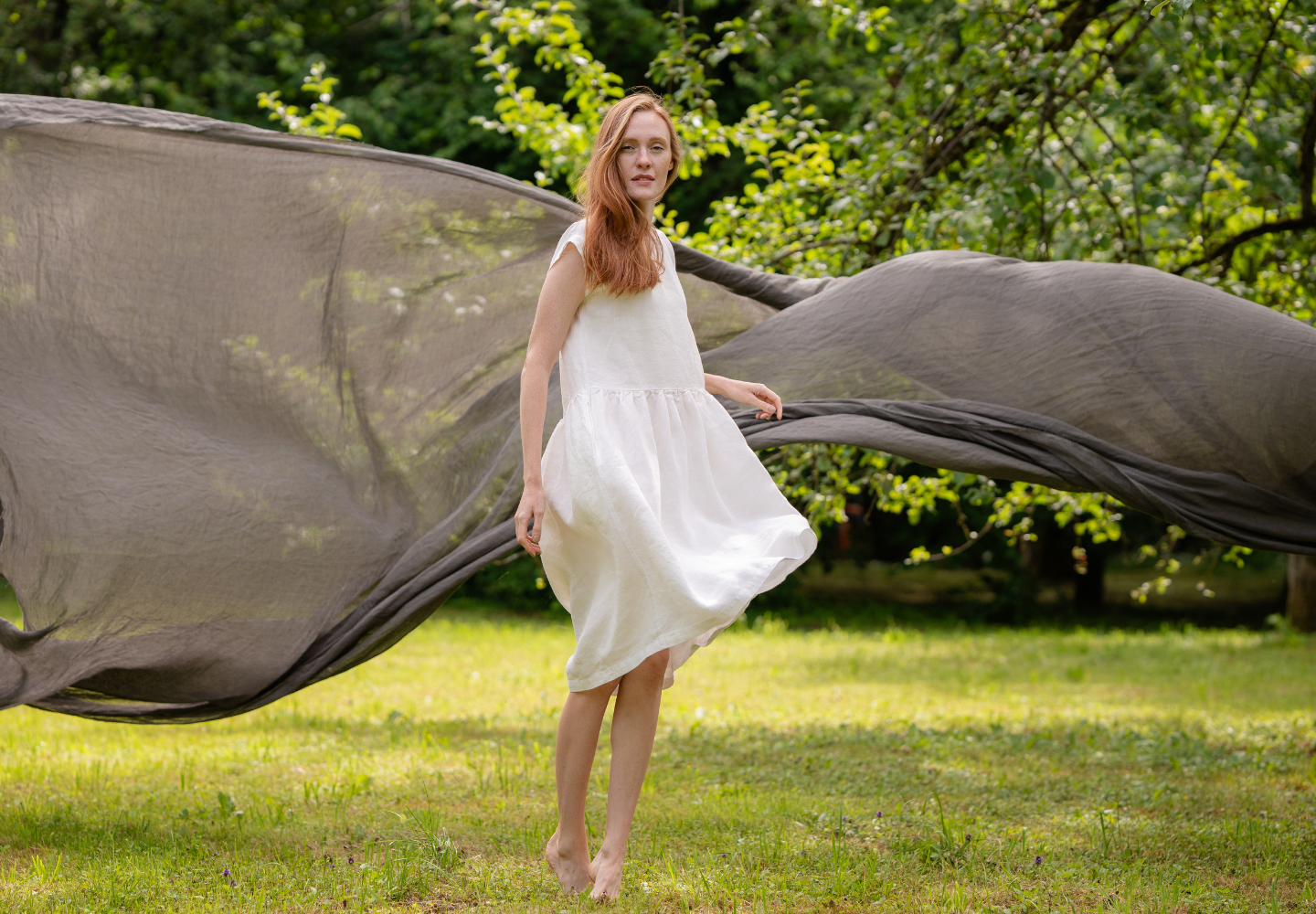 A woman wearing a white linen dress from menique stands barefoot on grass in a lush green garden, with a large flowing fabric creating a dynamic backdrop behind her.