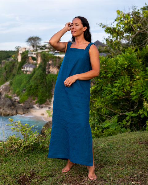 A woman with long dark hair, wearing a sleeveless, linen cobalt blue maxi dress with a square neckline and tie straps at the shoulders, stands barefoot on a grassy cliff overlooking a rocky coastline and ocean. She has one hand raised near her face and is looking out towards the water. The background features lush green vegetation and some buildings on the cliffs. The sky is overcast.