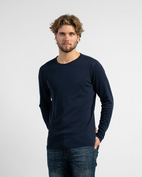 A three-quarter view of a man standing against a neutral studio backdrop, wearing a dark navy long-sleeve shirt paired with dark jeans.