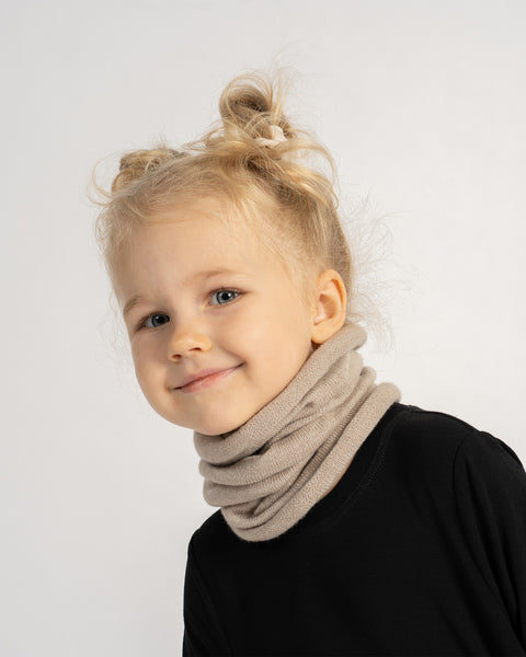 Little girl with blonde hair wearing a beige knit neck warmer and black outfit, smiling softly at the camera.