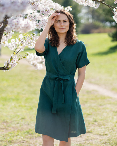 A woman with brown hair, wearing a dark green, short-sleeved wrap dress, stands outdoors under a tree with white blossoms.
