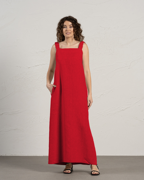 A woman with curly brown hair is standing in front of a textured off-white wall. She is wearing a menique sleeveless, floor-length pure red dress with wide straps and a square neckline. One of her hands is tucked into the side pocket of the dress. She is wearing black open-toe sandals and looking towards the right side of the frame with a slight smile. The lighting is even, casting soft shadows.