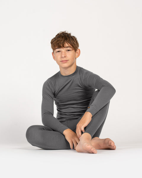 Older boy in gray Merino wool base layer set sitting cross-legged on the floor, neutral background.
