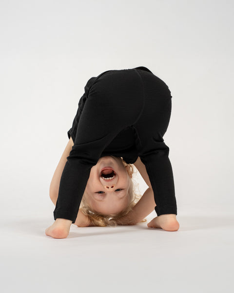 A young child with light-colored hair is playfully bent over, looking between their legs, against a plain white background. They are wearing dark-colored pants. The child's head is inverted, and they are smiling widely with their mouth open, looking directly at the camera. Their bare feet are visible on the floor.