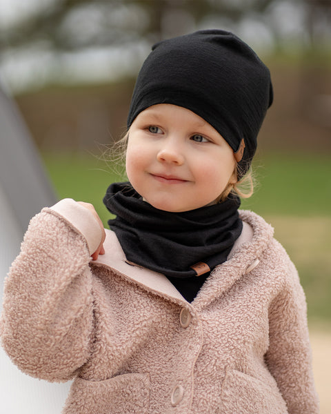 A young child with fair skin and light hair is looking slightly to the right with a gentle smile, wearing a menique black beanie and a matching black neck warmer. They are dressed in a cozy-looking, light pink or beige textured coat, with a blurred outdoor background.