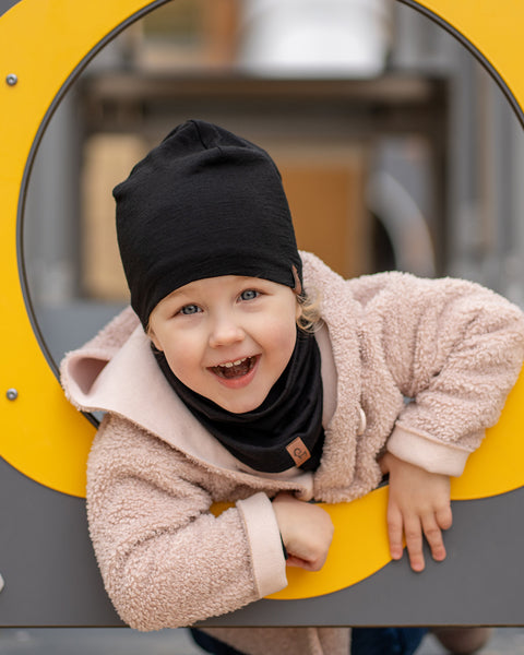 Smiling child playing outdoors wearing a black Merino wool kids’ beanie and matching neck gaiter, cozy beige coat, playground background.