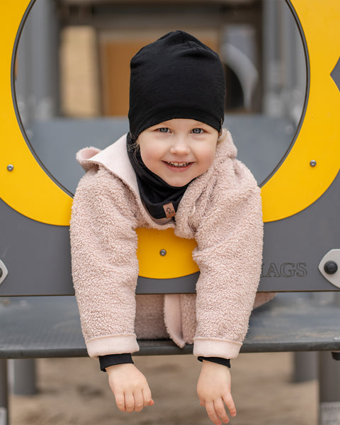 A young child with fair skin and light eyes is smiling broadly at the camera, peeking through a circular opening on a yellow and gray playground structure. They are wearing a menique black beanie, a black neck warmer, and a light pink or beige textured coat.