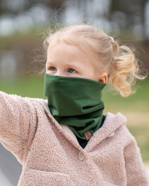 A young child with blonde hair tied in pigtails looks upwards and to the side, wearing a dark green Menique neck gaiter pulled up over their nose and mouth, paired with a beige hooded jacket. The child's blue eyes are visible above the neck gaiter, suggesting a moment of curiosity or observation outdoors.