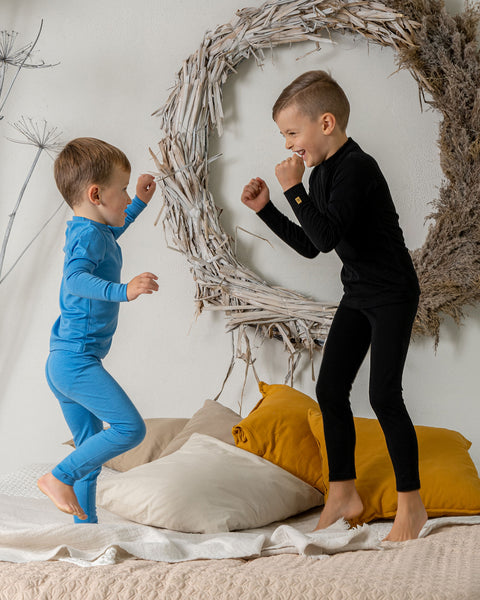 Two boys jumping on a bed, one of them is wearing the menique 160 kids' set of long sleeve crew and pants in light blue and the other in black color. They are play-fighting.