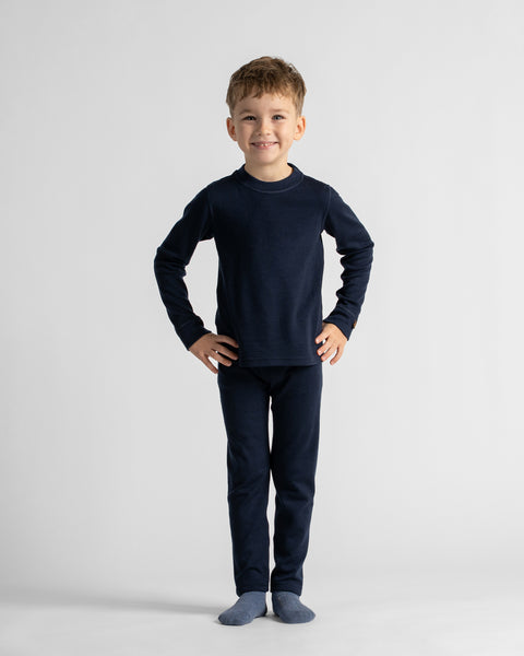 A full-length portrait of a young boy standing with hands on his hips against a plain backdrop, wearing a dark navy long-sleeve pajama or base layer set.