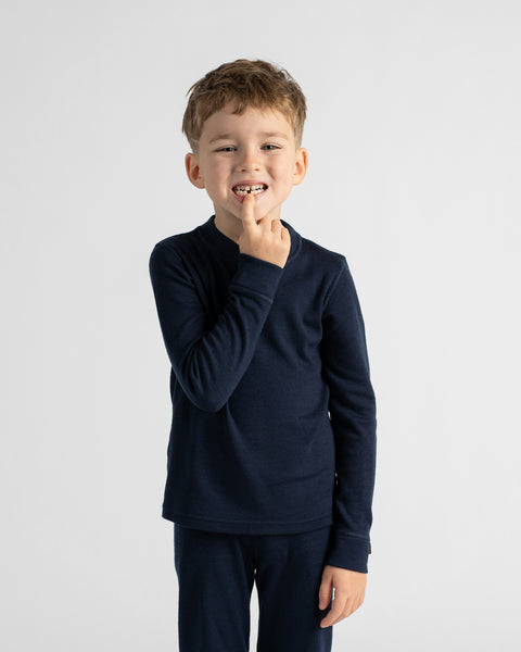 A young boy with short light brown hair stands against a light gray studio background, wearing a dark navy long-sleeve crewneck shirt and matching navy pants.
