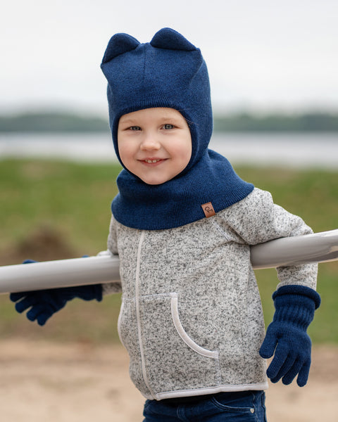 A young child with blue eyes and a slight smile, is leaning on a metal railing outdoors. They are wearing a dark blue balaclava with ears, dark blue gloves, and a grey speckled zip-up hoodie. The background is a blurred outdoor scene with green grass and water.