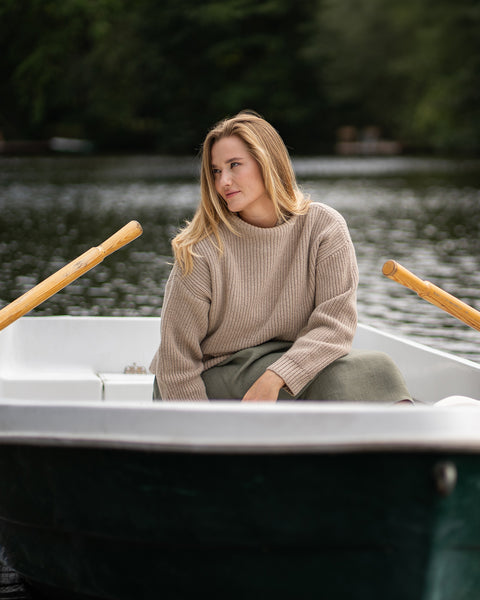 A woman wearing a beige knit sweater leans casually in a rowboat on the water. The image captures a quiet lakeside moment with a cozy, everyday outdoor style.