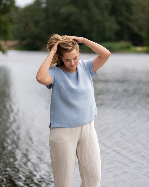 A barefoot woman leans gracefully against a wooden pier railing, gazing off to the side while brushing a hand through her hair. She wears a natural linen top and stone green bermuda shorts, with a peaceful lake stretching out behind her.