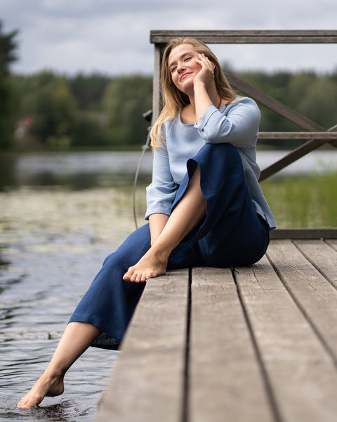 A woman relaxes on a wooden pier by a serene lake, dipping her bare foot into the water with her eyes closed and a peaceful smile. She is wearing a loose-fitting, cloudy blue long-sleeve linen top paired with wide-leg, storm blue linen pants