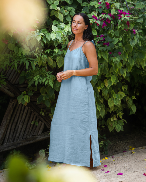 A light-skinned person with long dark hair is standing outdoors, looking slightly to the left with a serene expression. They are wearing a cloudy blue sleeveless maxi dress with a V-neck and side slits. Lush green foliage and vibrant pink flowers surround them, suggesting a tropical or garden setting.