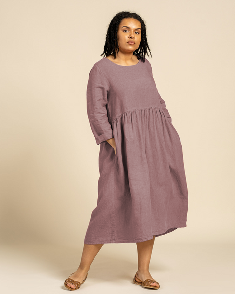 A woman with dark curly hair stands looking at the camera, wearing a menique linen long-sleeved dress in the color faded rose. Her hands are in the side pockets of the dress. She is positioned against a solid light brown background.