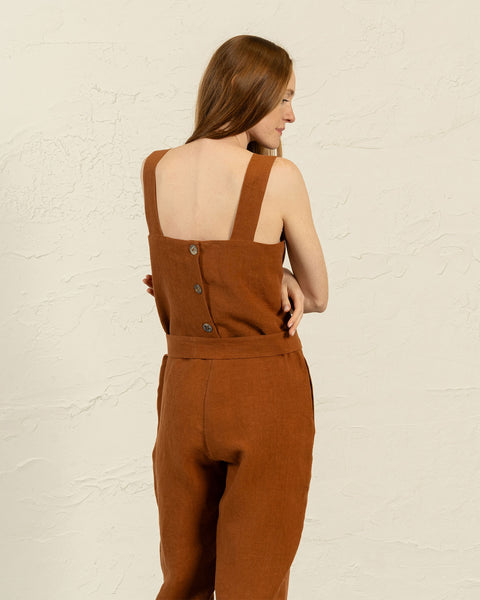 From the back, a person with long brown hair is shown wearing an almond brown colored jumpsuit that fastens with three buttons down the upper back. The jumpsuit also features a tied belt at the waist.