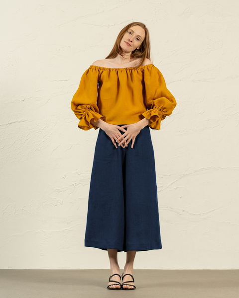 A light-skinned woman with long brown hair poses in front of a textured beige wall. She is wearing a spicy yellow, off-the-shoulder blouse with gathered neckline. Her bottoms are wide-legged, storm blue linen culottes that fall below her knees. She is barefoot and her hands are gently resting on her lower abdomen. She is looking slightly down and to her left with a soft expression. The lighting is even, casting minimal shadows.