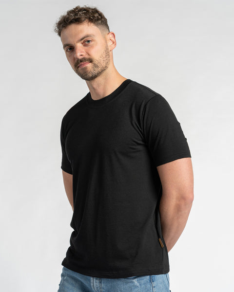 A man wearing a merino wool black t-shirt and blue jeans is shown from the chest up against a light background. He has short brown hair and a light beard, and is looking slightly to the side. The menique black t-shirt is the primary focus, showcasing its fit and appearance.