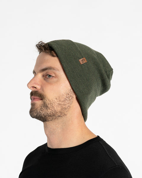 A person with light skin, short brown hair, and a beard is shown in profile against a white background. They are wearing a dark green knit beanie with a small brown tag on the side. They are also wearing a black t-shirt.
