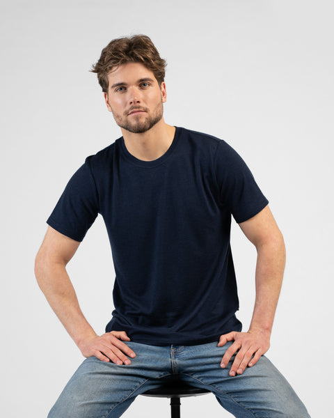 A studio portrait of a man sitting on a black stool against a plain white background. He is leaning slightly forward with his hands resting on his upper thighs, looking directly at the camera. He is wearing a dark navy blue short-sleeved crew-neck t-shirt and blue jeans.