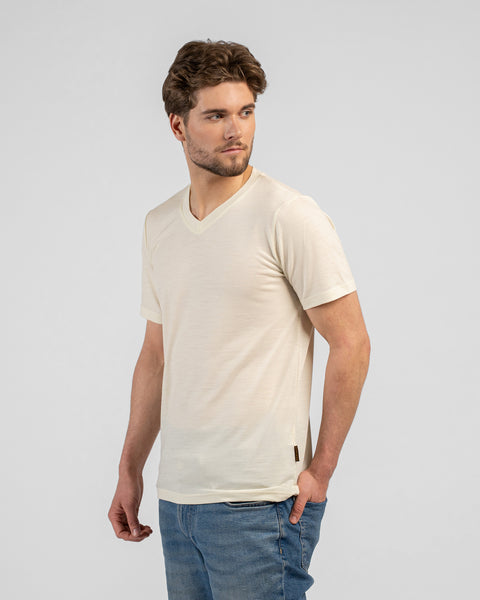 A man stands looking back over his right shoulder with his left hand resting in his pocket. He is wearing a light beige v-neck t-shirt and blue jeans against a light grey background.