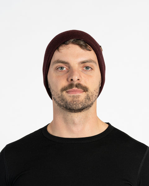A person with light skin, short brown hair, a mustache, and light-colored eyes is shown in a headshot against a white background. They are wearing a royal cherry knit beanie with a small brown tag on the side and a black long-sleeved shirt. They are looking directly at the camera.