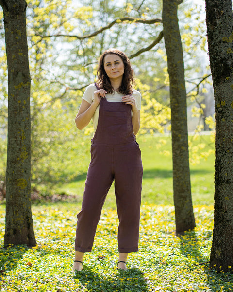 A woman with brown hair, wearing dark purple overalls over a light-colored t-shirt, stands outdoors between two tree trunks on grassy ground.