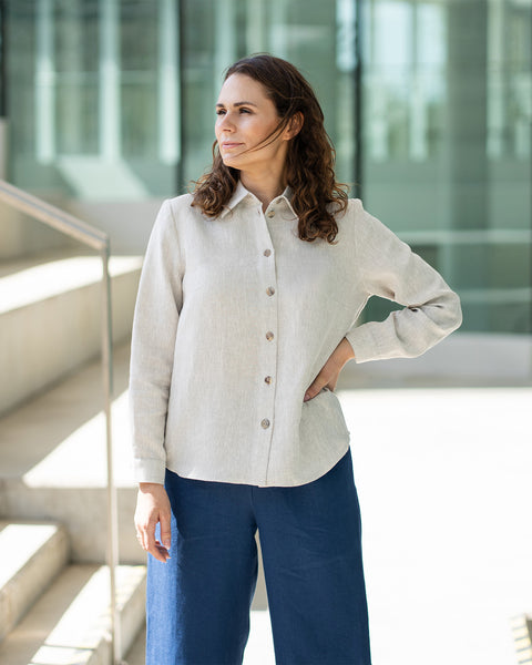 A woman with brown hair, wearing a natural linen button-front shirt and wide-leg white trousers, stands outdoors against a modern concrete structure.