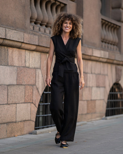 A smiling person with voluminous curly hair walks towards the camera, wearing a pure black menique sleeveless wrap top that ties at the waist and wide-legged black trousers, paired with black ballet-style shoes.
