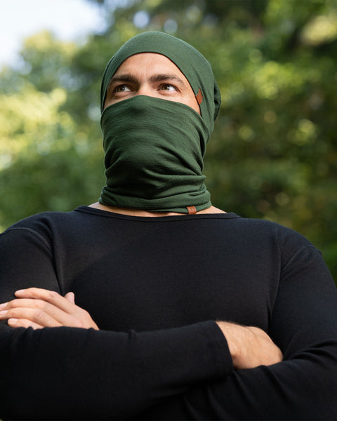 The picture features a man wearing the dark green unisex gaiter and beanie merino set. He has his arms crossed. He is in a forest.