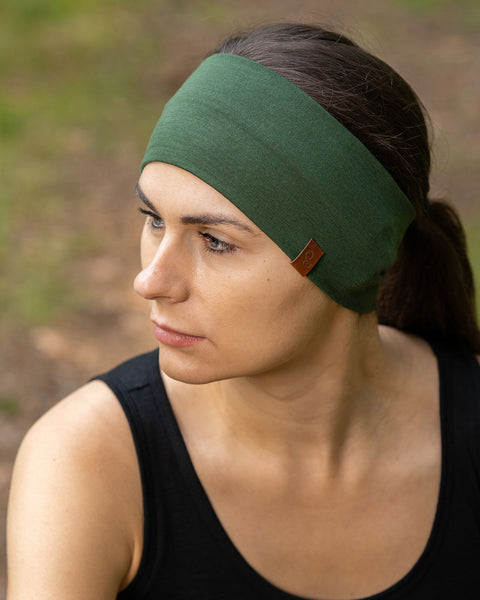 The picture features a woman sitting down in a forest, wearing the unisex dark green menique merino HEADBAND. She is also wearing a sleeveless black merino top. She is looking to the side.