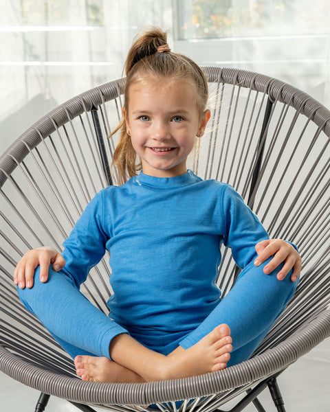 A girl sitting in a lounge chair wearing the menique Kids' 160 Merino Pants Light Blue color. Her legs are crossed and palms are on the knees.