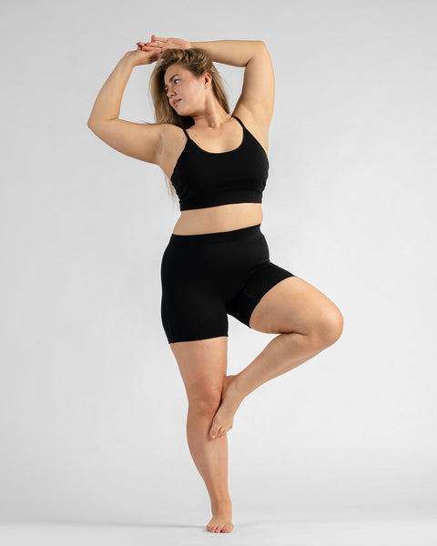 Front full-body view of a model standing on one leg with her hands resting behind her head, wearing a black sports bra with thin straps and black biker shorts.