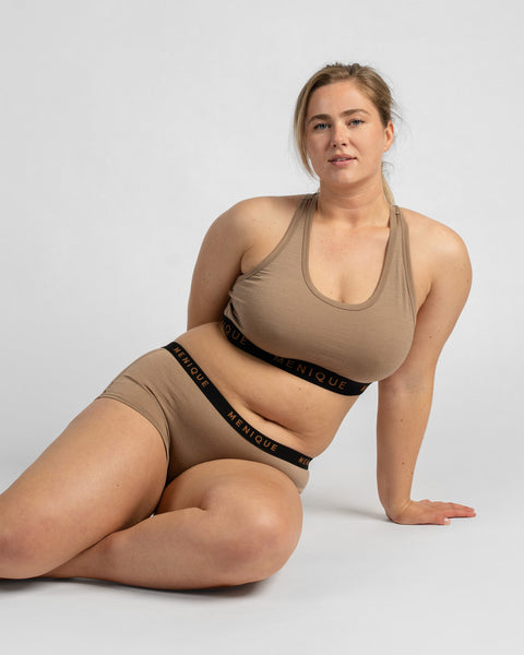 A woman sitting on the floor leaning back on one arm, wearing a beige sports bra and beige boxer briefs, both featuring a black elastic band with the word "MENIQUE".