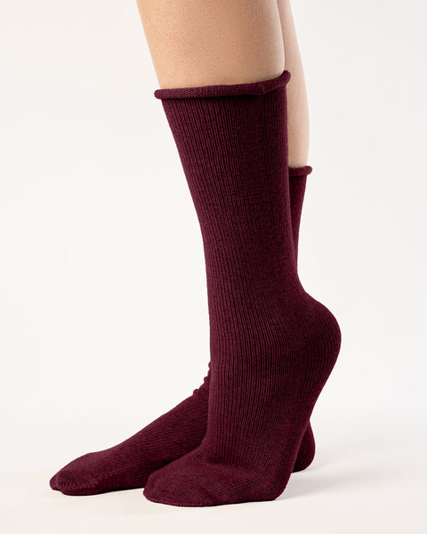 A side view shows a person from the mid-thigh down, wearing ribbed, menique red socks. The socks reach the mid-calf, and the person's feet are positioned with one slightly in front of the other, heel raised. They stand against a plain white background with even lighting that emphasizes the texture of the socks.