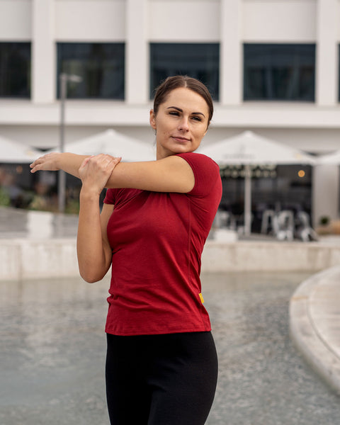 Woman in a merino wool menique t-shirt in the color royal cherry stretching outdoors with a building in the background