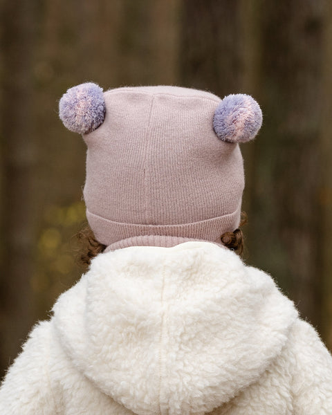 The back of a child's head and shoulders are shown, with the child wearing a light pink knit hat adorned with two pink and purple speckled pom-poms on top. They also wear a lighter pink turtleneck, and the hood of a cream-colored sherpa jacket is visible, set against a blurred forest background.