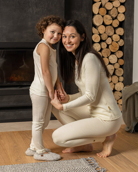 A woman is kneeling on a light-colored wooden floor, facing a child. Both are wearing matching merino wool long-sleeved tops and pants. The woman has long dark hair and is smiling.