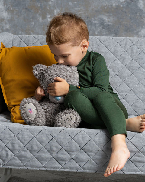 A young child with light brown hair, wearing a dark green long-sleeved shirt and matching pants, is sitting on a gray quilted sofa and hugging a gray teddy bear with a blue nose. The child's face is pressed against the bear.