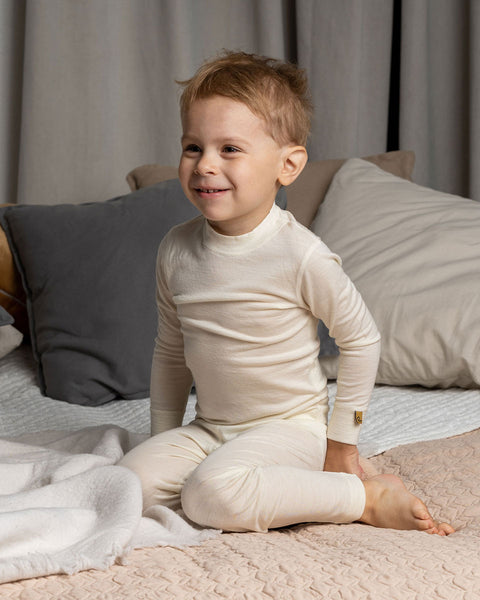 a boy sitting on a bed, wearing the Kids' Merino 160 Long Sleeve Set in Natural color.
