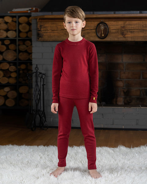 Young boy wearing a matching royal cherry thermal base layer set, standing barefoot on a white rug in front of a fireplace with stacked firewood in the background.