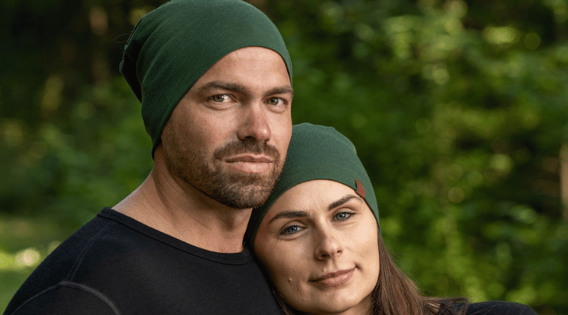 Two people, a couple, wearfing matching 160gsm dark green merino beanie hats in a forest.