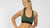 A young, blond woman wearing a dark green merino wool sport bra with menique logo band. She has one hand on her hip, and is smiling while looking to the side.