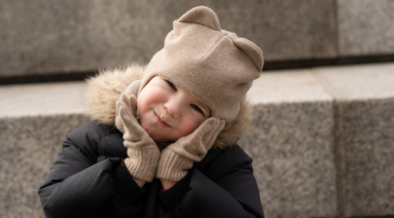 a toddler girl holding her cheeks in her palm, wearing a beige balaclava with ears and matching mittens.