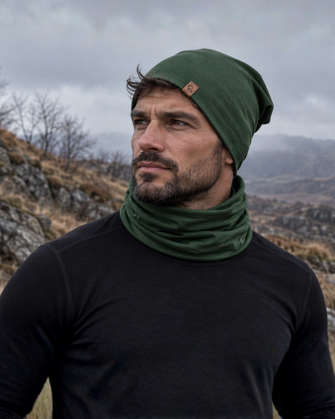 A bearded man wearing a dark green beanie and a matching neck gaiter looks thoughtfully off to the side. He is dressed in a black long-sleeved thermal top, set against a rugged, mountainous landscape under a heavily clouded sky.