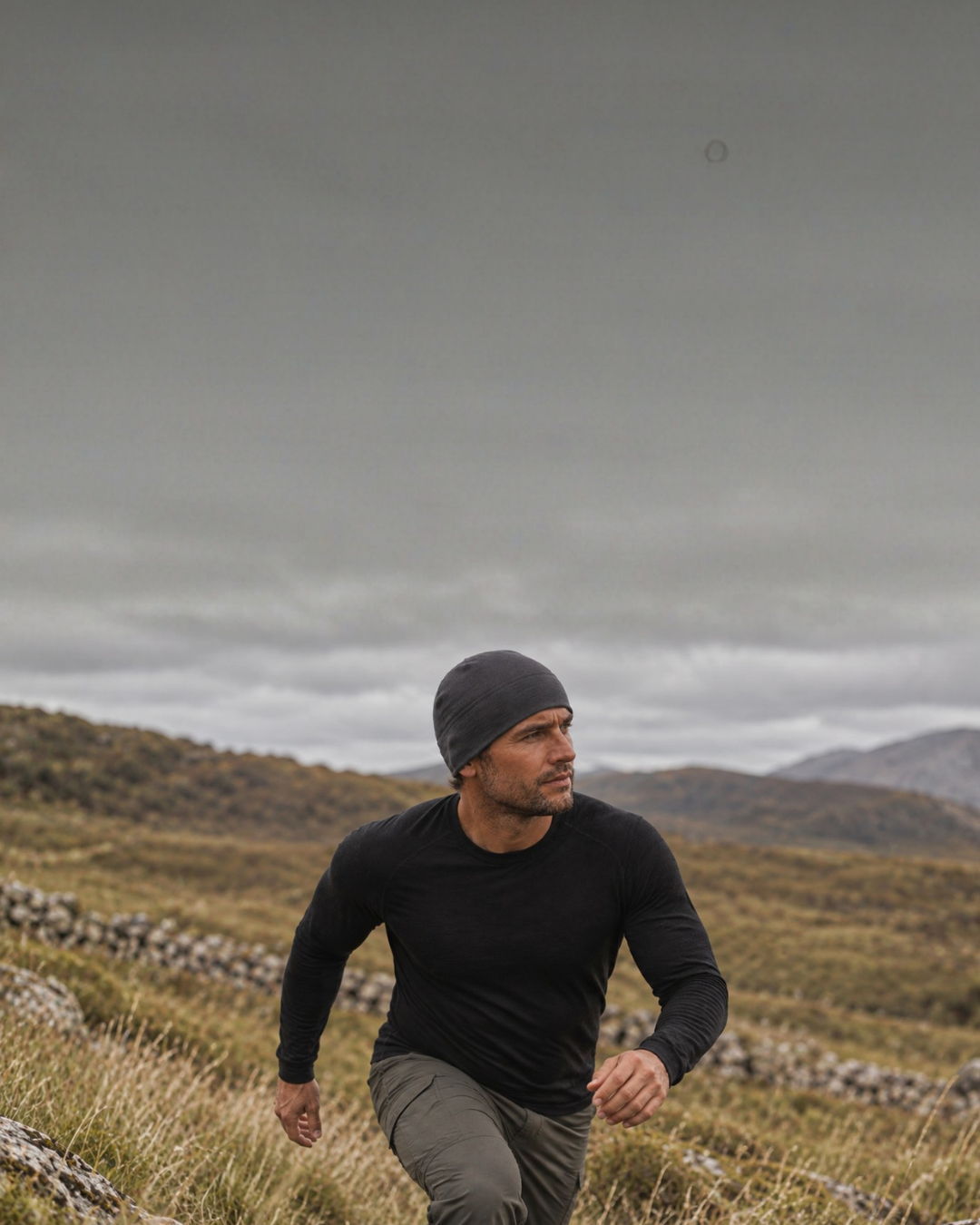 A man wearing a black long-sleeved thermal top and a dark grey beanie hikes up a rugged, grassy hillside. He looks intently off to the side, set against a vast backdrop of rolling mountains and a dark, moody overcast sky.