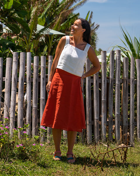 A woman wearing a white top and an cinnamon red skirt stands in front of a bamboo fence, looking up and smiling.